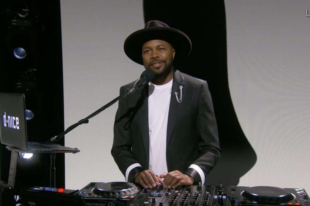 DJ D-Nice in a suit standing behind his DJ tech set up
