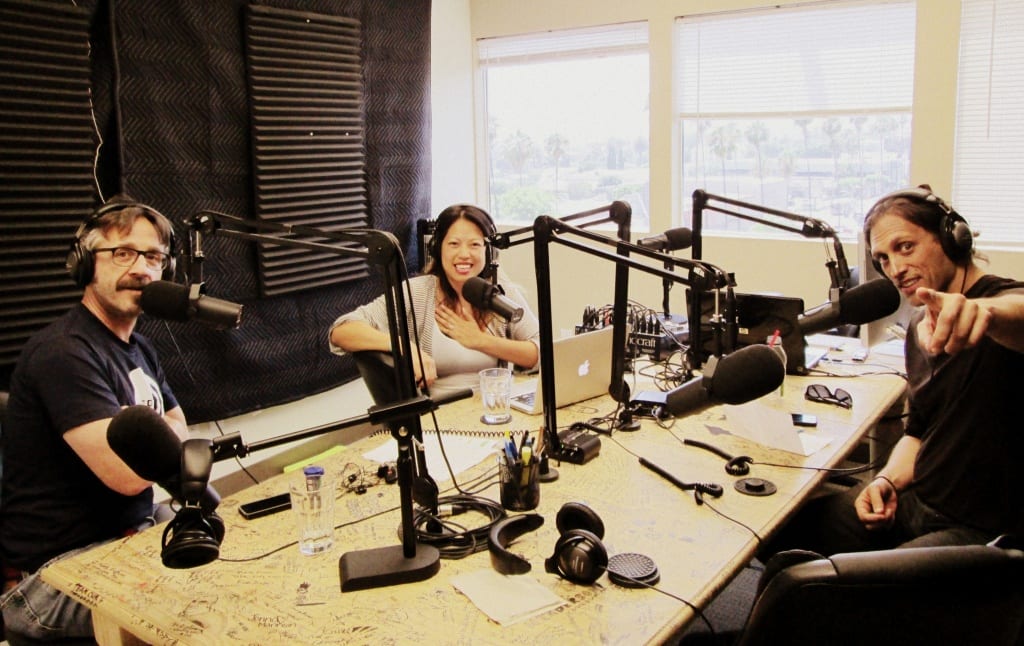 Photo of a woman and two men sitting at a table in front of microphones inside a recording space.