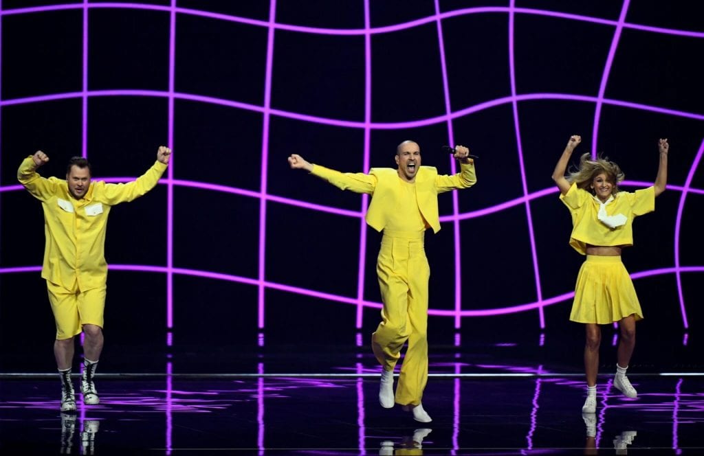 Three people in bright yellow dancing against a black and purple checkered background onstage