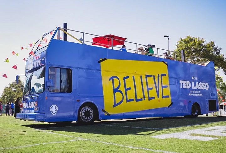 Blue Ted Lasso promotion bus with large yellow "BELIVE" sign on the side