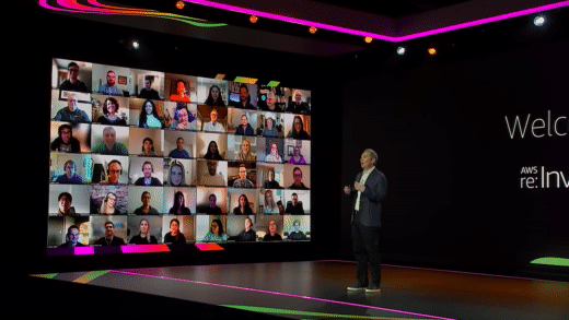 Man giving an in person presentation on stage with virtual attendees displayed on the wall beside him
