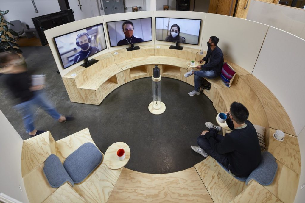 Two men sitting in a hybrid meeting space, walking to 3 people on virtual screens